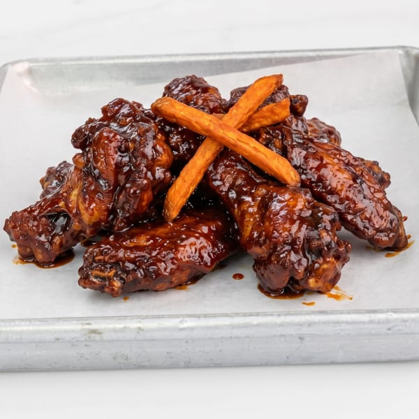 Dark, saucy chicken wings with sweet potato fries on a metal tray with white paper.