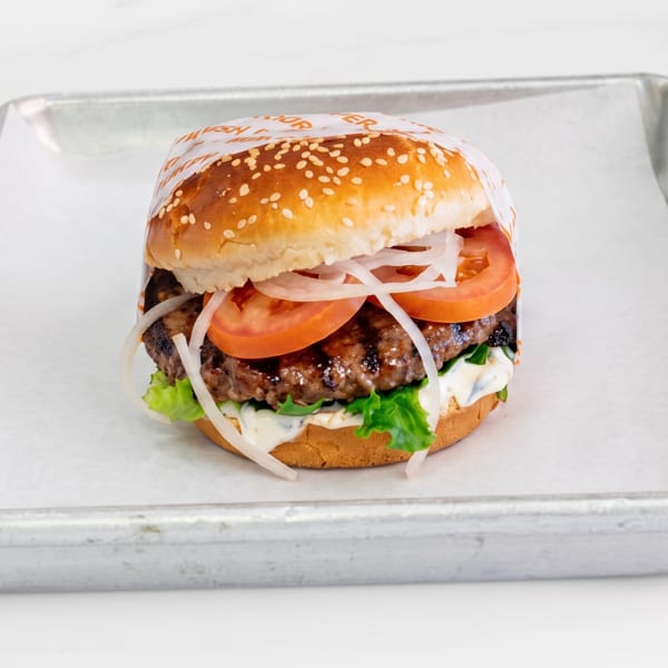 Grilled burger with lettuce, tomatoes, onions, and white sauce on a sesame bun, partially wrapped on a tray.