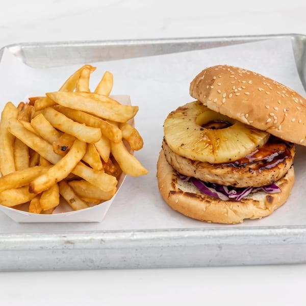 A grilled chicken and pineapple burger with red cabbage slaw and a side of french fries on a metal tray.